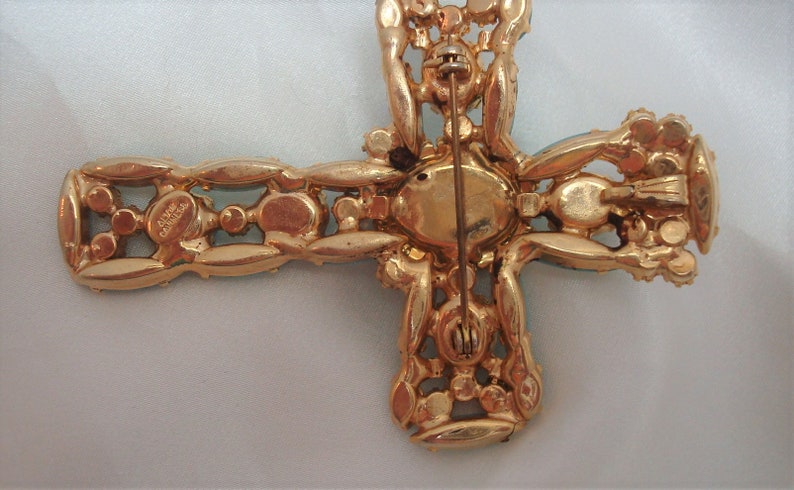 May include: Gold tone cross brooch with clear and blue rhinestones. The back of the brooch has a pin and a signature that reads "Alice Caviness".