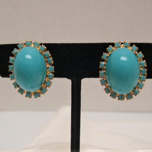 May include: A pair of turquoise blue clip-on earrings with gold-toned settings. Each earring features a round turquoise stone surrounded by a halo of smaller gold-toned stones.
