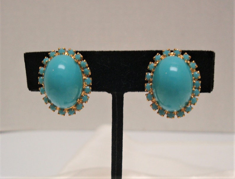 May include: A pair of turquoise blue clip-on earrings with gold-toned settings. Each earring features a round turquoise stone surrounded by a halo of smaller gold-toned stones.