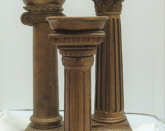 May include: Three gold-coloured decorative candle holders shaped like columns with ornate details. The candle holders are arranged in a row on a white surface.