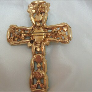 May include: A gold-tone cross brooch with clear and green rhinestones. The back of the brooch has a pin and a signature.