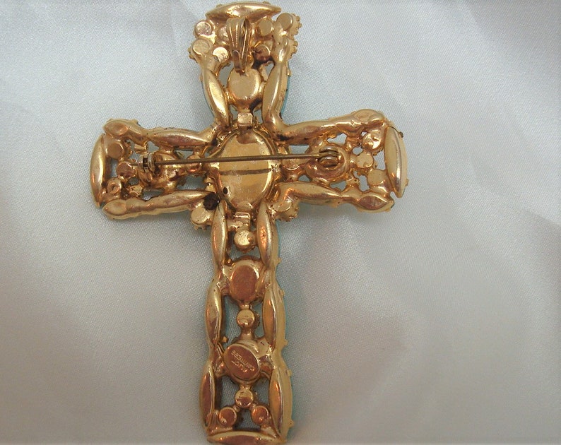 May include: A gold-tone cross brooch with clear and green rhinestones. The back of the brooch has a pin and a signature.