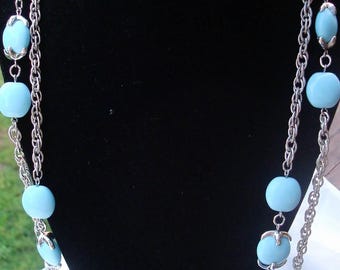 May include: A multi-strand silver-tone chain necklace featuring light blue, smooth, rounded beads. The beads are interspersed along the metallic chains and are secured by small silver-tone metal prongs that grip each individual bead.