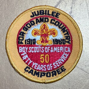 May include: A gold and red embroidered patch with the text "JUBILEE FOR GOD AND COUNTRY 1910 1960 BOY SCOUTS OF AMERICA FIFTY YEARS OF SERVICE CAMPOREE" and a red and gold fleur-de-lis.
