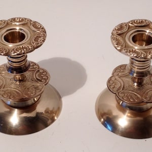 May include: A pair of gold metal candle holders with ornate designs. Each holder has a round base, a fluted column, and a decorative top with a central opening for a candle.