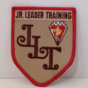 Puede incluir: Un parche bordado de color beige y rojo con el texto "JR. LEADER TRAINING" y las letras "LT" en marrón. Un diamante rojo con un contorno blanco y una flor de lis amarilla en su interior está por encima de las letras.