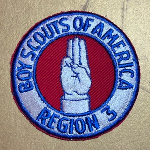 May include: A white and blue embroidered patch with the text "Boy Scouts of America Region 3" in a circle. The centre of the patch features a white hand making the Boy Scout sign.