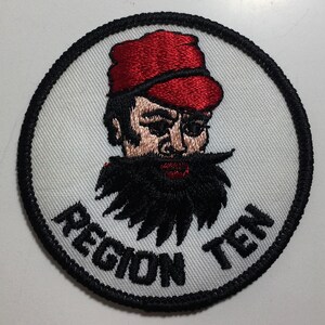 May include: A white embroidered patch with a black border. The patch features a cartoon man with a red hat and a black beard. The text "REGION TEN" is written in black below the man.