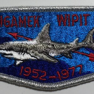 Puede incluir: Un parche bordado azul y blanco con un tiburón, flechas y el texto "AMANGAMEK WIPIT 470 WWW BSA 1952-1977".