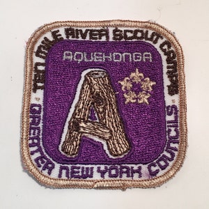 Puede incluir: Un parche bordado de color morado y marrón con el texto "Tet Mile River Scout Council Greater New York Council" y "Aquehonga" en blanco. El parche presenta una letra "A" estilizada hecha de dos troncos marrones.