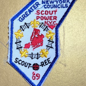 May include: A white embroidered patch with a blue border. The patch features a red and yellow graphic of a city skyline with the text "GREATER NEW YORK COUNCILS SCOUT POWER NYC" and "SCOUT-REE 69".