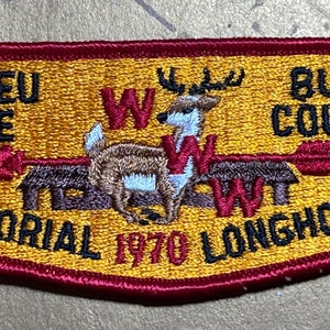 Puede incluir: Un parche bordado amarillo y rojo con un ciervo de cola blanca, una cabaña y flechas. El texto dice "Ajapeu Lodge 33 Memorial 1970 Longhouse Bucks County PA."