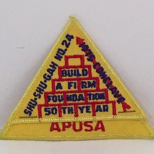 Puede incluir: Un parche triangular amarillo con texto bordado en azul y rojo. El texto dice "SHU-SHU-GAH NO. 24 1945 CONCOURSE BUILD A FIRM FOUNDATION 50TH YEAR APUSA".