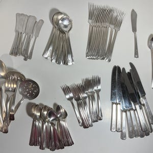 May include: A collection of vintage silverware, including forks, spoons, and knives, in a variety of styles. The silverware is arranged in groups, with some pieces overlapping. The silverware is all silver in color and has a polished finish.