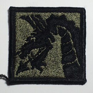 Puede incluir: Un parche bordado negro y verde con un diseño de dragón. El dragón está mirando hacia la izquierda y tiene una cola larga y puntiaguda. El parche está rodeado por un borde negro.