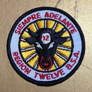 May include: A white and black embroidered patch with a bull's head in the centre. The bull's head is black with a red nose and a white circle with the number 12 in the centre. The patch is surrounded by a white circle with the text "SIEMPRE ADELANTE REGION TWELVE BSA" in red and black.