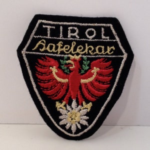 Puede incluir: Un parche bordado negro con las palabras "TIROL" y "Safelekar" en blanco. El parche presenta un águila roja con detalles dorados y una flor de edelweiss blanca.