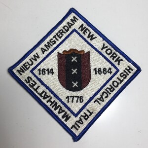 May include: A diamond-shaped patch with a blue border and white background. The patch features text that reads "NIEUW AMSTERDAM NEW YORK HISTORICAL TRAIL MANHATTES" and the years 1614, 1664, and 1776. A shield with three white crosses is in the center.