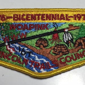 Puede incluir: Un parche bordado amarillo, verde y azul con el texto "1776 Bicentenario 1976" y "Lincoln Trails Council". El parche presenta un río, árboles y un símbolo nativo americano.