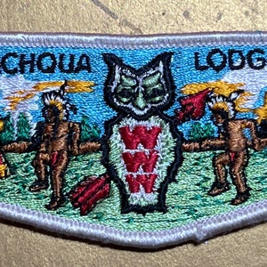 Puede incluir: Parche bordado con el texto "RANACHQUA LODGE NO 4". El parche presenta un búho con símbolos "W", figuras nativas americanas y un paisaje con árboles y rayos. El parche tiene un borde blanco.