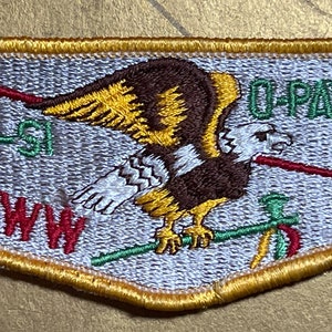 May include: A white, gray, and green embroidered patch with a brown eagle with yellow wings and a red arrow in its beak. The patch has the text "O-PAW-GAN 1162" and "MI-GI-SI WWW" in green and red.