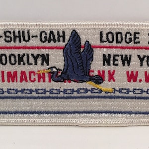 Puede incluir: Parche blanco bordado con detalles azules y amarillos. El parche presenta una garza azul volando con una flecha roja apuntando hacia la izquierda y el texto "SHU-SHU-GAH LODGE 24 NEW YORK WIMACHINK W.W. BROOKLYN".