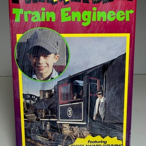 May include: A colorful box with the text "I WANNA BE A Train Engineer" in yellow and green. It features a photo of a smiling child in a train engineer hat and a vintage train. The box also includes the text "Featuring Emmy Award Winning Host Steve Pool".