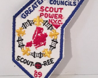 May include: A white embroidered patch with a blue border. The patch features a red and white map of New York City, surrounded by a wreath of gold fleur-de-lis. The text "GREATER NEW YORK COUNCILS SCOUT POWER NYC." is above the map, and the text "SCOUT-REE 69" is below the map.