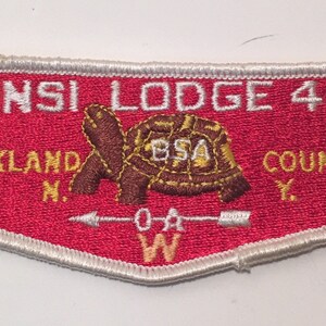 Op de afbeelding: Een geborduurde rode en witte patch met de tekst "Munsi Lodge 444 W Rockland N.Y. County W" en een bruine schildpad met de tekst "BSA" op zijn schild. De schildpad kijkt naar rechts en er is een witte pijl die naar rechts wijst met de tekst "OA W" eronder.