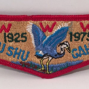 Puede incluir: Un parche bordado de color marrón claro y rojo con una garza azul de pie en el agua. El parche tiene el texto "W 1925 SHU SHU W 1975 GAH-24".