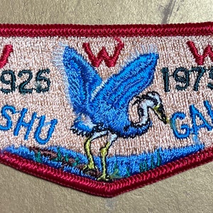以下が含まれることがあります： 青いサギのデザイン、テキスト「SHU SHU」と「GAH-24」、1925年と1975年が刺繍されたパッチ。パッチは、赤いボーダーと赤い「W」の文字が付いた、薄いタン色の背景です。