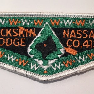 Puede incluir: Un parche bordado verde y blanco con el texto "BUCKSKIN LODGE NASSAU CO. 412" y una silueta de lobo negro dentro de un pino verde.