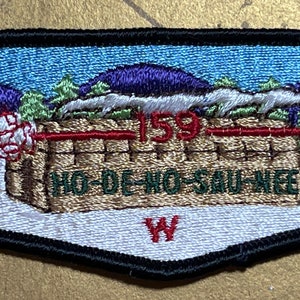 Op de afbeelding: Een vintage geborduurde patch met een winterscène. De patch toont een hut met een rode pijl die naar rechts wijst, een besneeuwd landschap en de tekst "HO-DE-NO-SAU-NEE" en "159".