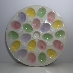 May include: A round white ceramic egg serving platter featuring eighteen oval indentations in pastel shades of yellow, green, pink, and purple. The tray includes embossed chick and bunny figures between the egg cups and the text HAPPY EASTER.