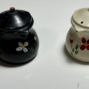 May include: Two small ceramic salt and pepper shakers shaped like pots with lids. The shaker on the left is black with white flowers and the shaker on the right is white with red flowers.