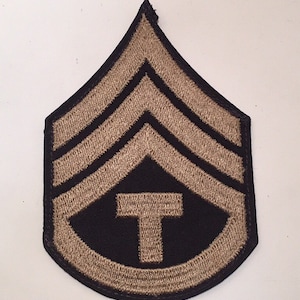 May include: A tan embroidered military patch with three chevrons and a 'T' in the centre on a black background.