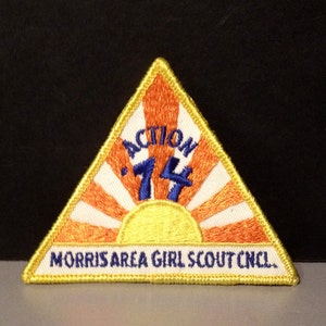Puede incluir: Un parche triangular amarillo con fondo blanco y rayos naranjas. El parche tiene texto azul que dice "ACTION 74" y "MORRIS AREA GIRL SCOUT CNCL."