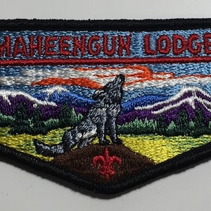 Puede incluir: Un parche bordado azul y verde con un lobo aullando a la luna. El parche dice "Maheengun Lodge" y tiene una flecha roja apuntando hacia la izquierda. El parche también tiene las letras "W" repetidas tres veces.