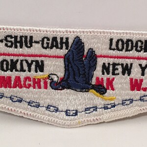 Puede incluir: Parche blanco bordado con texto rojo y negro que dice "SHU-SHU-GAH LODGE 24 BROOKLYN NEW YORK WIMACHT NK W.W.". Un pájaro azul con pico y patas amarillas vuela a través del parche con una flecha roja apuntando hacia la derecha.