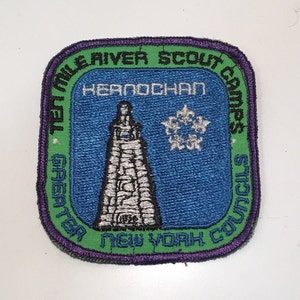 Puede incluir: Un parche bordado en verde, morado y azul con el texto "Ten Mile River Scout Camps Greater New York Councils" y las palabras "Henochan" en una pancarta blanca. El parche presenta un copo de nieve blanco y una torre gris y blanca.