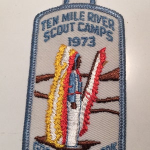 May include: A blue and white embroidered patch with the text "Ten Mile River Scout Camps 1973 Greater New York Councils BSA". The patch features a Native American figure in a feathered headdress.