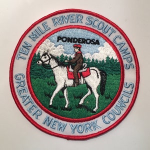 Puede incluir: Un parche bordado blanco con un borde rojo. El parche presenta un caballo blanco con un jinete, árboles verdes y nubes azules. El texto en el parche dice "Ten Mile River Scout Camps Greater New York Councils Ponderosa".