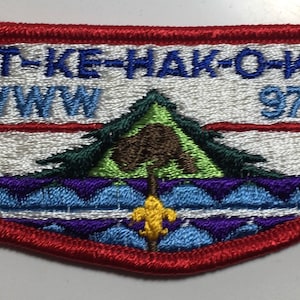 May include: A white embroidered patch with red trim. The patch features the text "KIT-KE-HAK-O-KUT WWW 97" and a stylized image of a pine tree with a blue arrow pointing to the right.