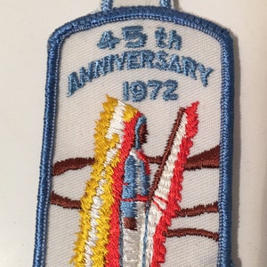 May include: A blue and white embroidered patch with the text "45th Anniversary 1972" and "T.M.P."  The patch features a stylised image of a person standing in front of a flag.