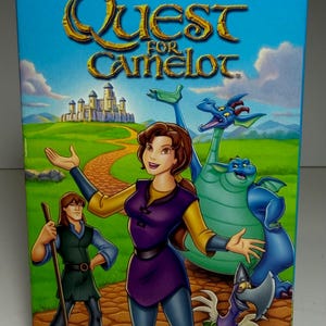 May include: VHS tape for the animated film "Quest for Camelot." The cover features cartoon characters, including a woman in purple and yellow, dragons, and a man with a staff. The title is in gold lettering against a blue sky.