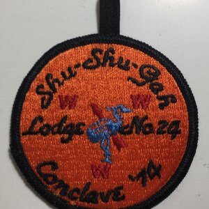 Puede incluir: Un parche redondo bordado de color naranja con costuras negras. El parche presenta el texto "Shu-Shu-Gah Lodge No. 29 Conclave '74" en letras cursivas negras. Un pájaro azul con alas rojas se encuentra en el centro del parche.