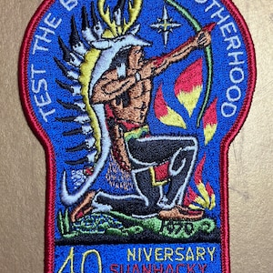 Può includere: Una toppa ricamata blu con bordo rosso. La toppa presenta un'immagine di un indigeno inginocchiato e che mira con arco e frecce. Il testo "Test the Bow of Brotherhood" è scritto in un cerchio attorno all'immagine. Il testo "40th Anniversary Suanhacky Lodge 49" è scritto sotto l'immagine.