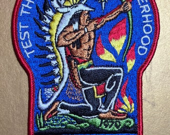 May include: A blue embroidered patch with a red border. The patch features an image of an indigenous person kneeling and aiming a bow and arrow. The text "Test the Bow of Brotherhood" is written in a circle around the image. The text "40th Anniversary Suanhacky Lodge 49" is written below the image.