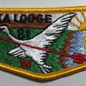 Puede incluir: Un parche bordado amarillo, verde, marrón, blanco y azul con el texto "Taleka Lodge 81" y un pájaro blanco con una flecha roja que lo atraviesa. El parche tiene un borde amarillo y una forma curva.