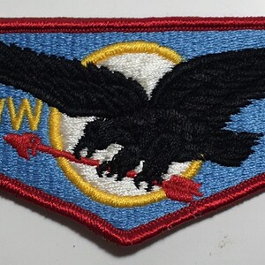 Puede incluir: Un parche bordado azul y rojo con un águila negra volando con una flecha en sus garras. El parche tiene el texto "WWW" y "94" en amarillo.